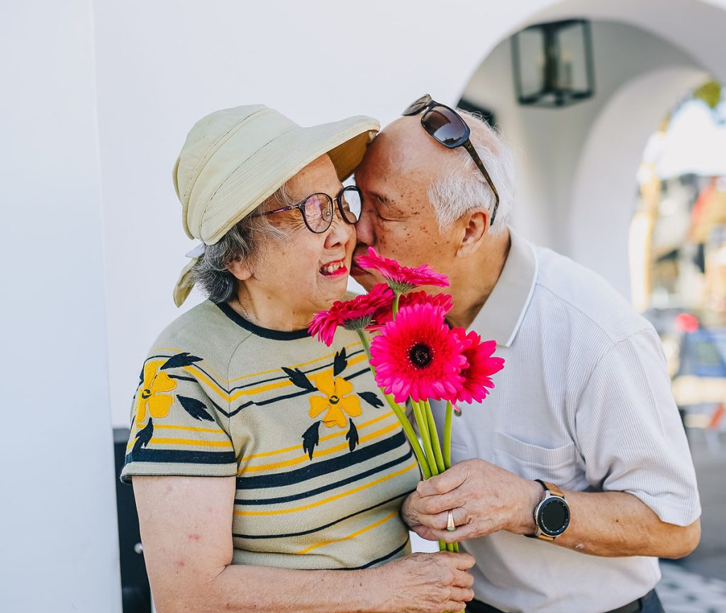 Elderly man kisses his wife on the cheek while holding pink gerberas