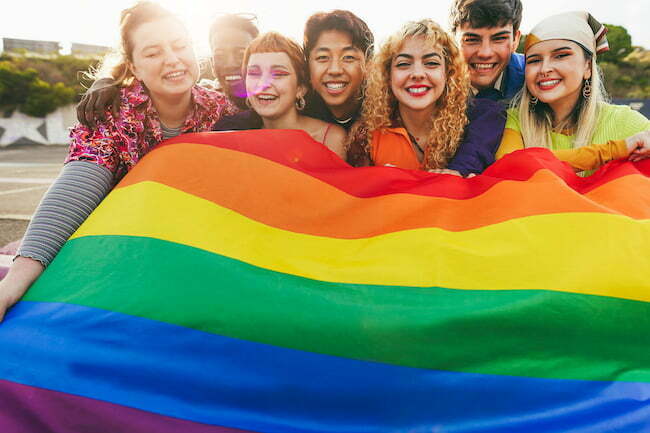 Young diverse people having fun holding LGBT rainbow flag outdoor - Focus on center blond girl